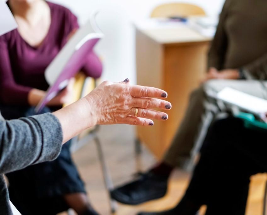 Hand shown in the foreground, gesturing with an air of instruction, before a group of seated people.